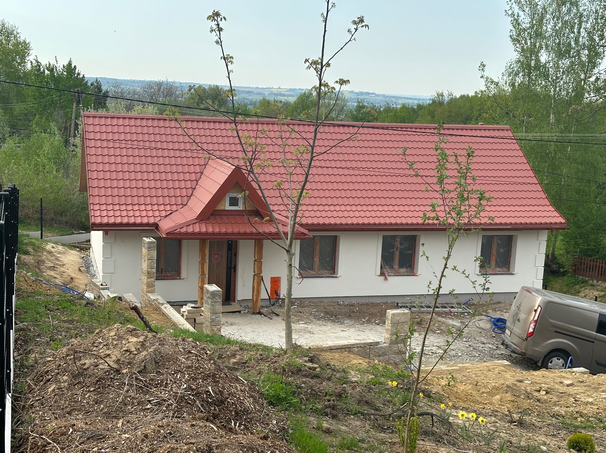 Dom z czerwoną dachówką w trakcie wykończenia, widoczny z góry. Częściowo zagospodarowany teren wokół, zaparkowany samochód dostawczy.