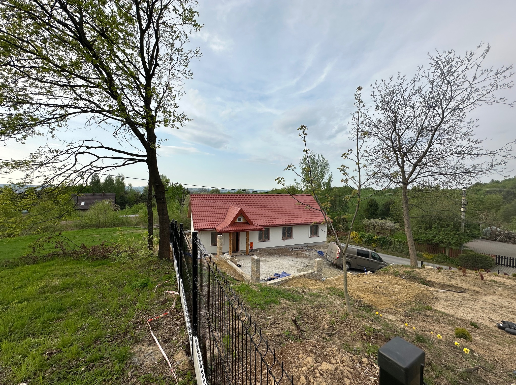 Dom jednorodzinny z czerwonym dachem w trakcie wykończenia, widok z góry, ogrodzenie panelowe na pierwszym planie, drzewa po bokach, krajobraz w tle.