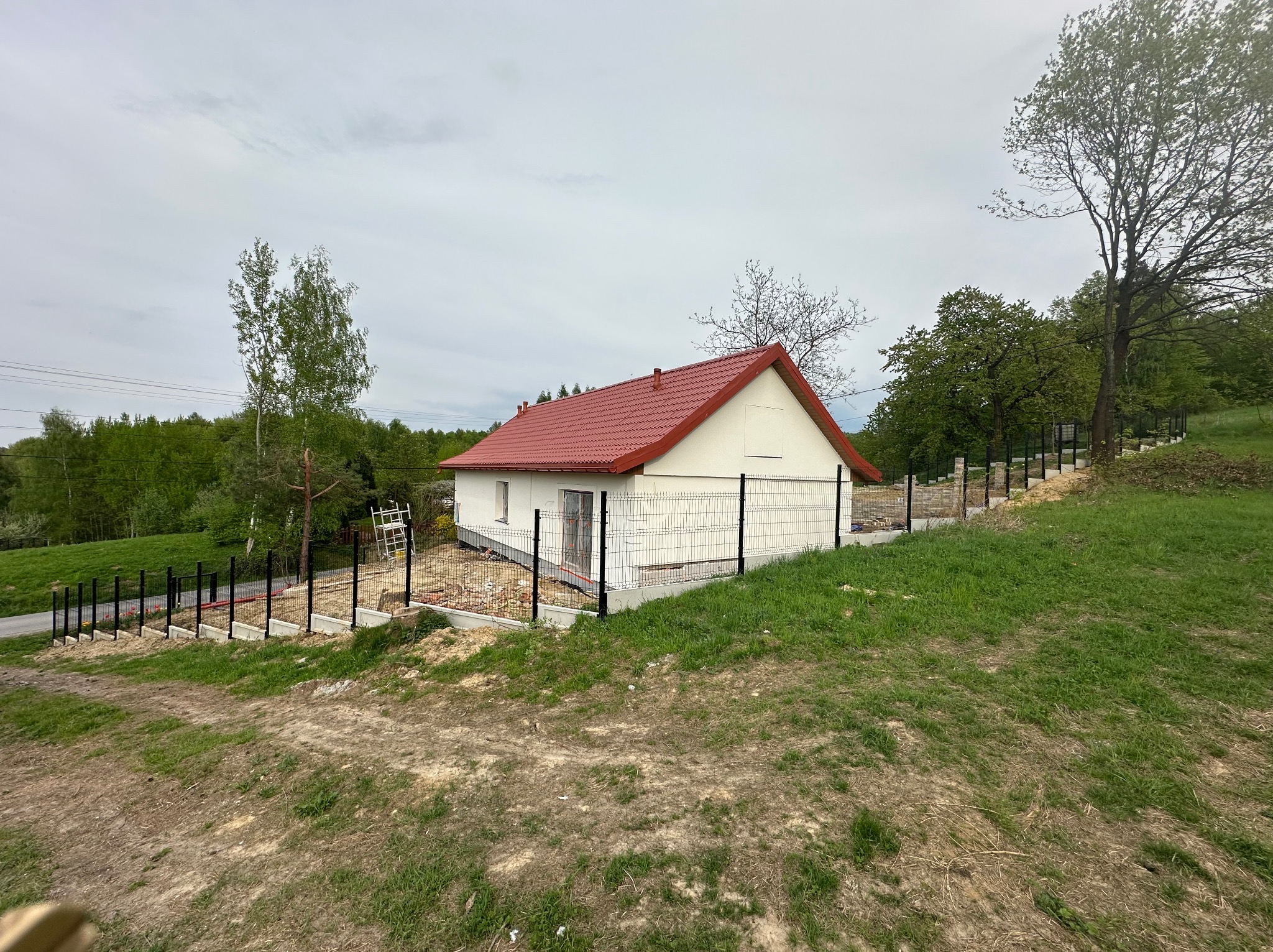 Nieduży, parterowy dom z czerwonym dachem, otoczony czarnym ogrodzeniem panelowym na nierównym terenie. Widoczne drzewa i zieleń w tle. Dom w trakcie wykończenia.