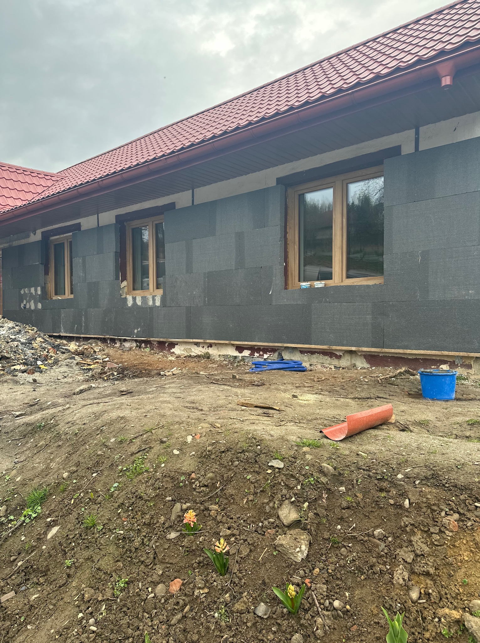 Ocieplanie elewacji grafitowym styropianem w Sułowie. Widoczne okna i fragment dachu z czerwonej dachówki. Prace w toku, teren wokół budynku częściowo uporządkowany.