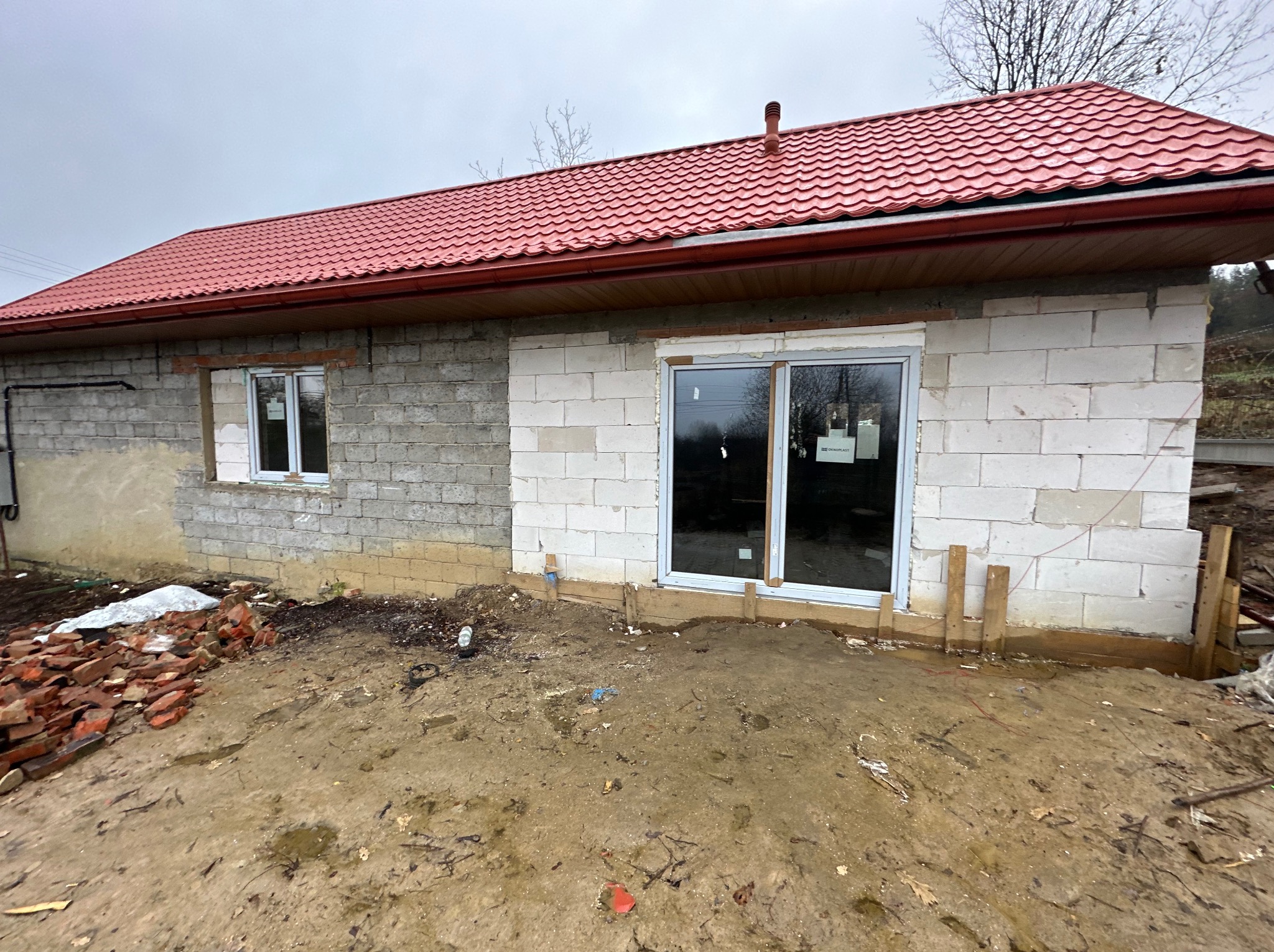 Budynek w trakcie budowy z czerwonym dachem, widoczne okna i drzwi, częściowo otynkowane ściany, gruz i błoto na zewnątrz, pochmurne niebo.