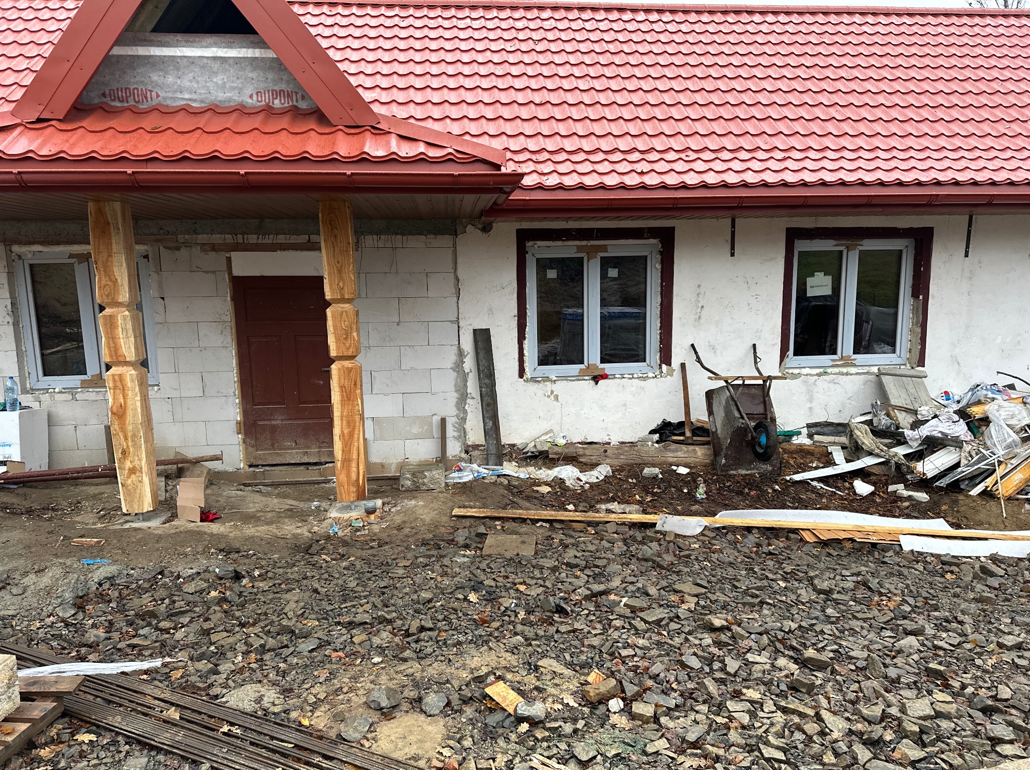 Dom w trakcie budowy z czerwonym dachem, widoczne okna, drewniane kolumny i gruz na podwórku. Ściany z białego bloczka.