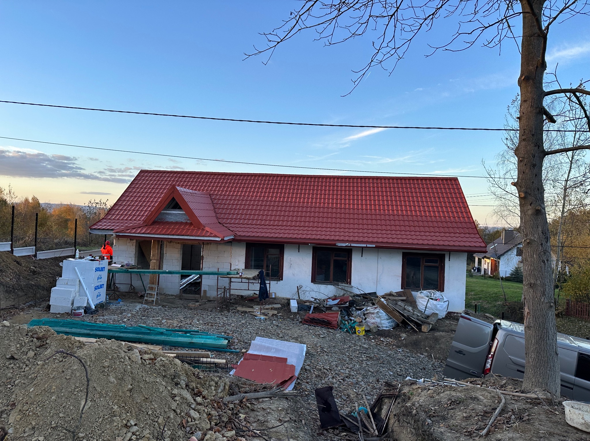 Dom w trakcie budowy z czerwonym dachem, widoczne materiały budowlane i częściowo ocieplone ściany. W tle drzewa i błękitne niebo.