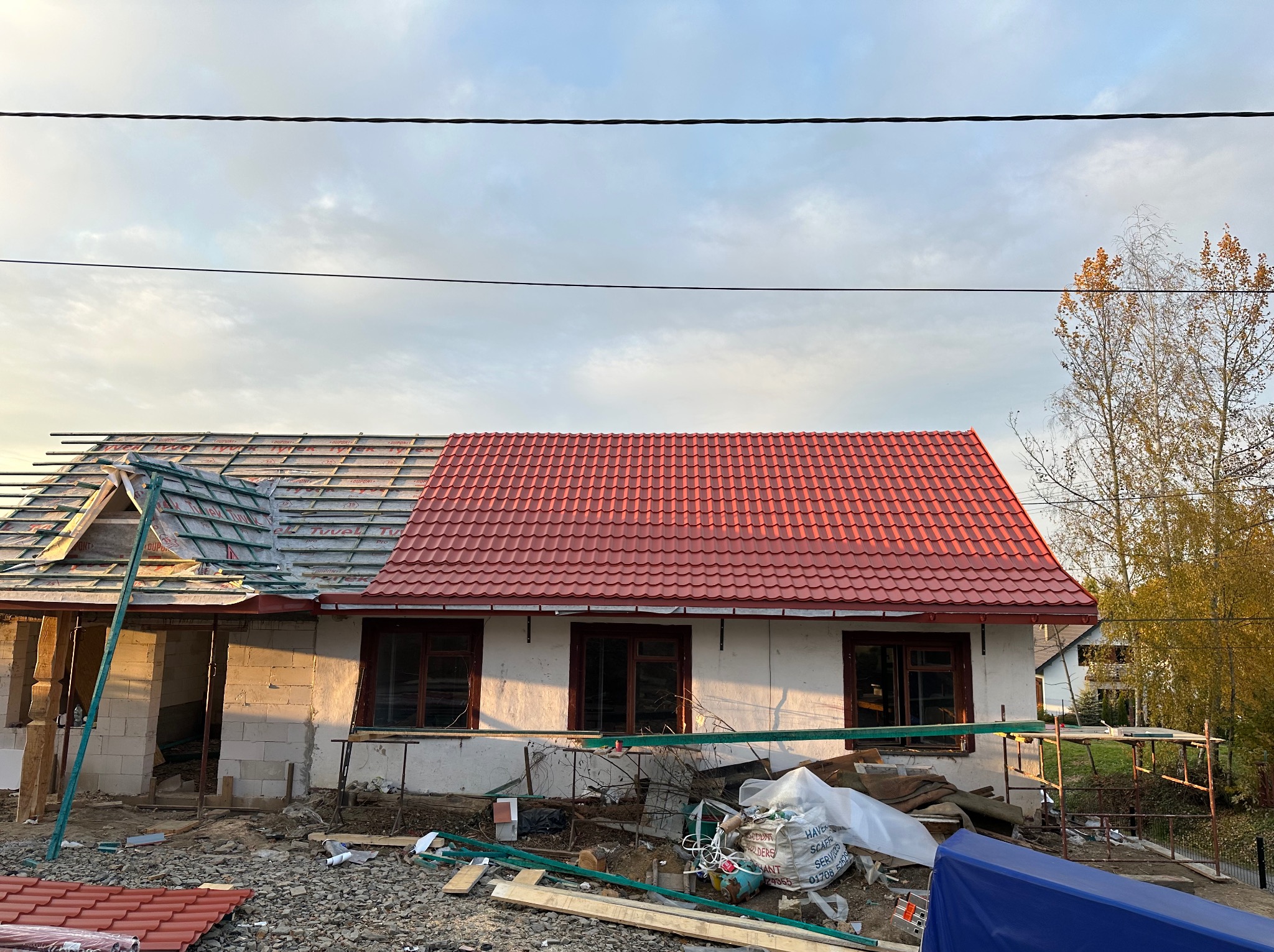 Dom w trakcie budowy z częściowo ułożonym czerwonym dachem. Widoczne materiały budowlane i rusztowania. Część dachu pokryta membraną dachową.