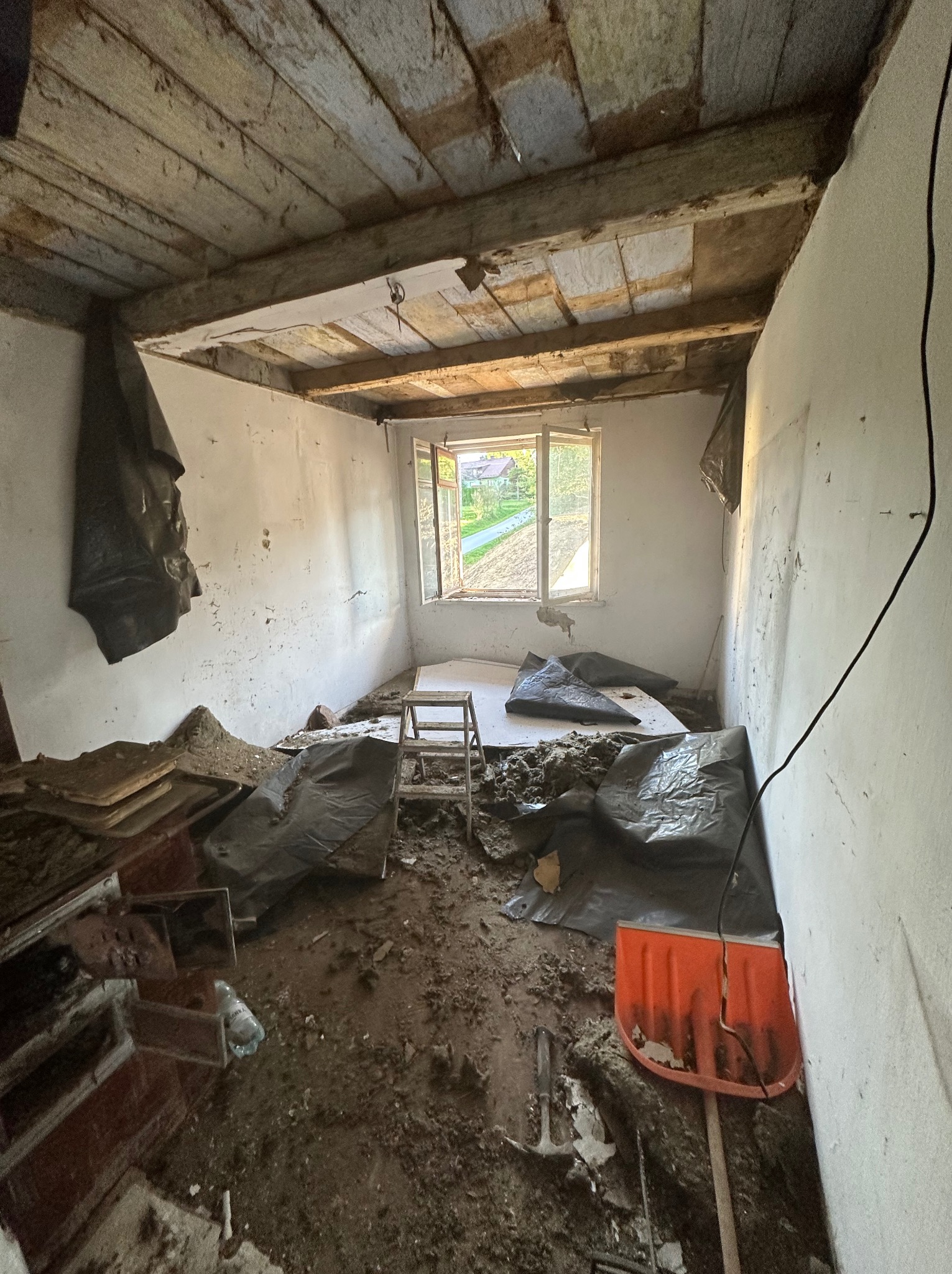 Wnętrze pokoju w trakcie remontu: widoczne stare drewniane deski na suficie, okno z widokiem na zieleń, gruz, folia ochronna i drabina. Przygotowanie do prac wykończeniowych.