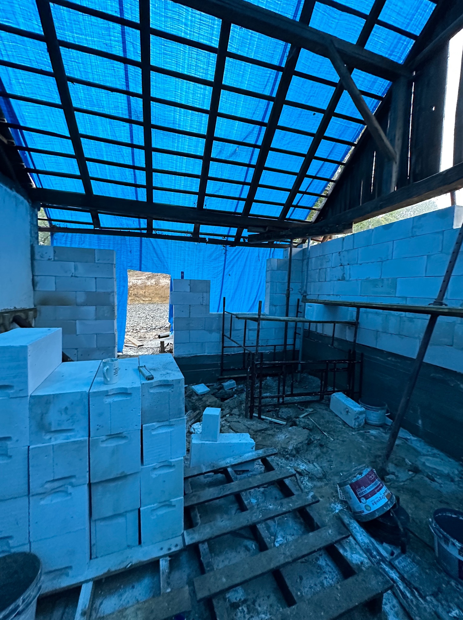 Wnętrze budynku w trakcie murowania ścian z bloczków, z widocznym niebieskim zadaszeniem i elementami rusztowania. Praca w toku, widoczne materiały budowlane.
