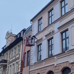 MALAR STUDIO - Pracownik na podnośniku koszowym maluje elewację kamienicy z ozdobnymi elementami architektonicznymi, widoczne zabezpieczenia okien folią.