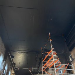 MALAR STUDIO - Wnętrze w trakcie prac: pomalowany na czarno sufit z widocznymi instalacjami, rusztowanie z osłoną i fragment ściany z oknami zabezpieczonej folią.