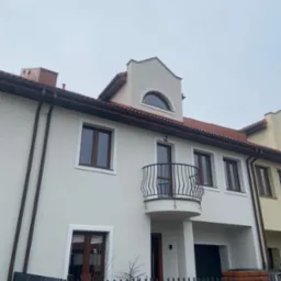 Jasna elewacja odnowionego segmentu z balkonem z czarną, kutą balustradą, brązowe okna i rynny, numer domu na ceglanym słupku przy furtce.
