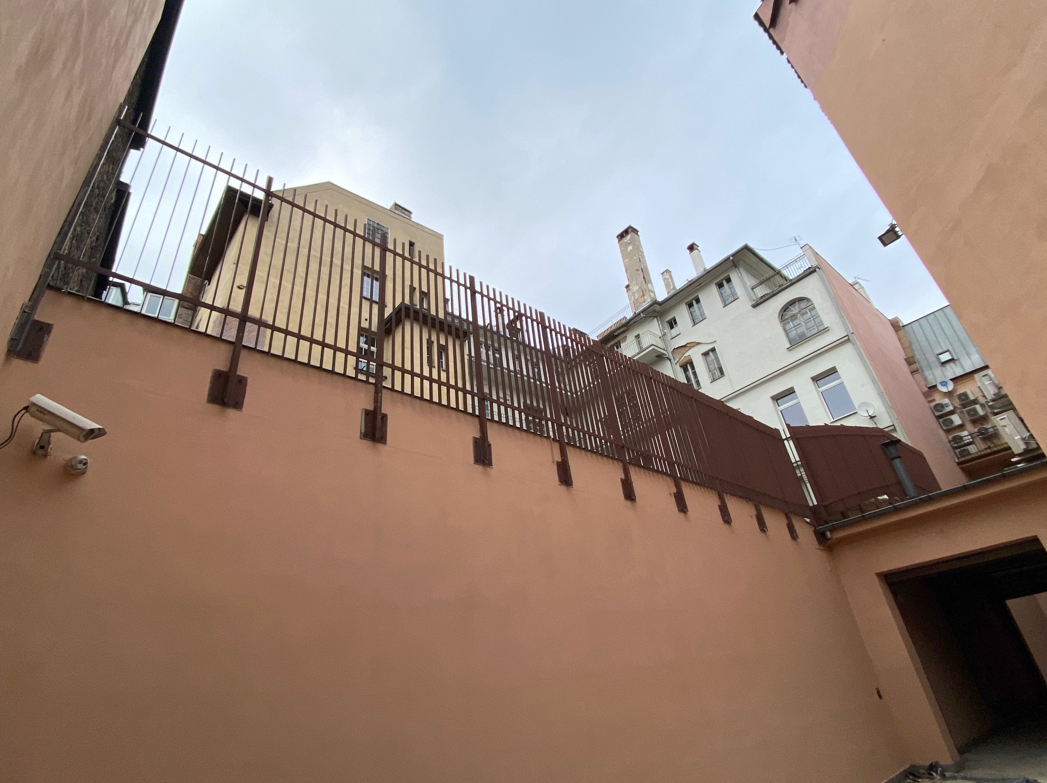 Widok z dołu na pomalowaną na różowo ścianę budynku z zamontowaną kamerą monitoringu i metalowym ogrodzeniem na górze, z widocznymi fragmentami innych budynków i pochmurnym niebem w tle.