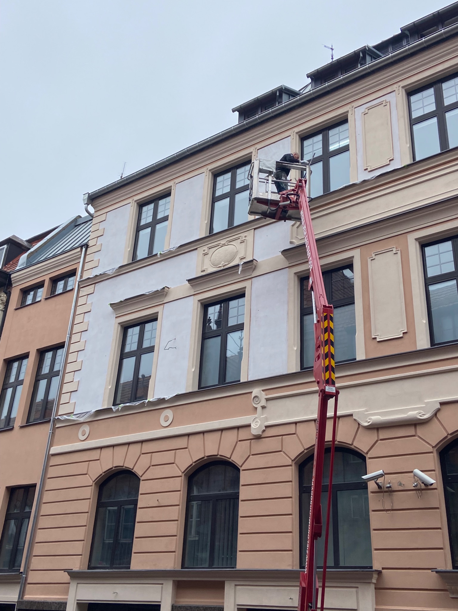 Pracownik na podnośniku koszowym podczas renowacji elewacji kamienicy, widoczne fragmenty odnowionej i przygotowanej do odnowienia powierzchni.