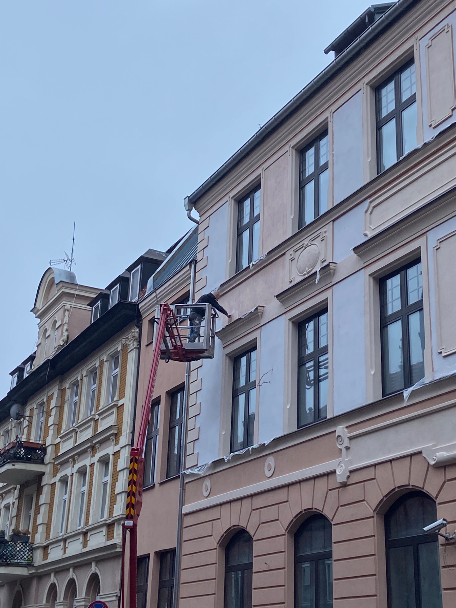 Pracownik na podnośniku koszowym maluje elewację kamienicy z ozdobnymi elementami architektonicznymi, widoczne zabezpieczenia okien folią.