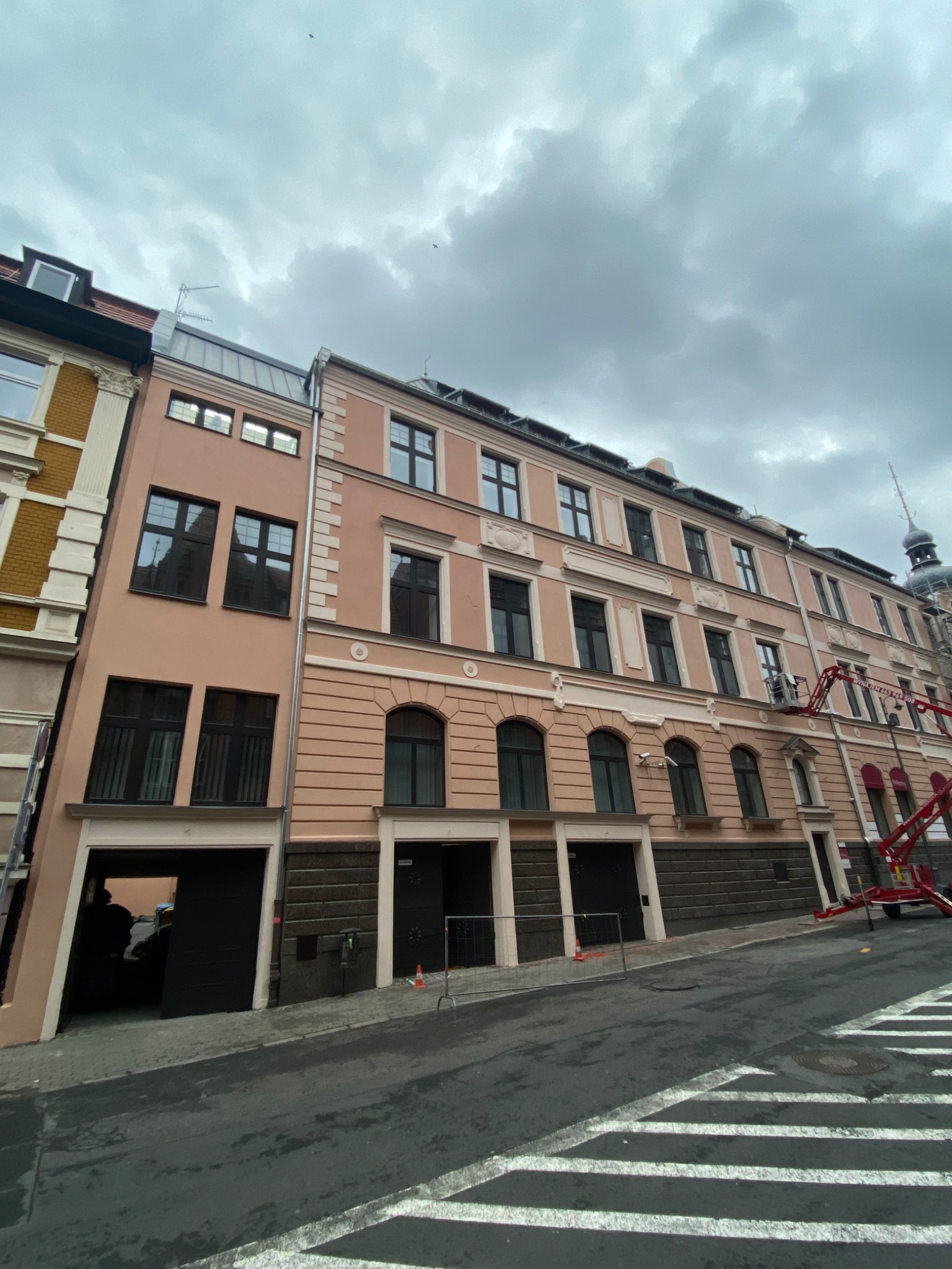Wielopiętrowy budynek w trakcie renowacji, z widoczną platformą roboczą na wysięgniku i odświeżoną, różową elewacją. Na pierwszym planie mokra ulica z pasami dla pieszych.