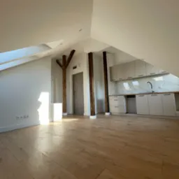 Przestronne, jasne wnętrze poddasza z drewnianymi belkami konstrukcyjnymi, nowymi panelami podłogowymi i zabudową kuchenną w odcieniach bieli, widok na okno dachowe