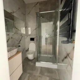 Nowoczesna łazienka z marmurowymi płytkami, kabiną prysznicową ze szklanymi drzwiami, umywalką w kształcie muszli na drewnianej szafce z dekoracyjnymi żłobieniami i czarnym grzejnikiem drabinkowym.