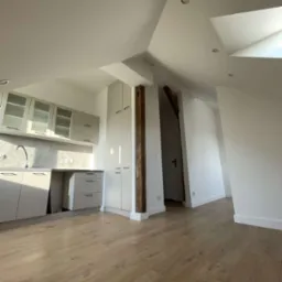 Jasna kuchnia w skosach poddasza z drewnianą podłogą, widok na fragment korytarza z drewnianą belką.