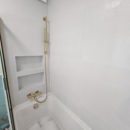 Michał Winiarski - Biała łazienka z wanną wypełnioną pianą, złota armatura, wnęka w ścianie wyłożonej płytkami. Minimalistyczny design, czyste linie, nowoczesne wykończenie.