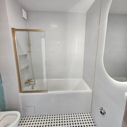 Michał Winiarski - Wykończona łazienka w szarościach z wanną i kabiną prysznicową w złotej ramie. Na podłodze biało-czarna mozaika. Owalne lustro z podświetleniem LED.