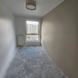 Michał Winiarski - Pusty pokój po gładziowaniu ścian, z widokiem na okno i grzejnik. Podłoga surowa, przygotowana do dalszych prac wykończeniowych. Ściany w odcieniach bieli i beżu.