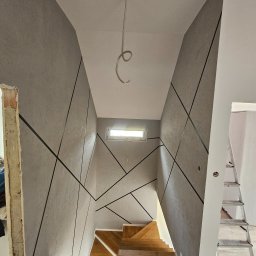 Michał Winiarski - Klatka schodowa po remoncie: szare ściany z czarnymi, geometrycznymi wzorami, drewniane schody, widoczny fragment niedokończonego otworu drzwiowego i drabina.