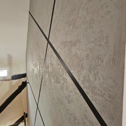 Michał Winiarski - Dekoracyjna ściana z szarą, teksturowaną gładzią i czarnymi, ukośnymi pasami. W tle widoczne rusztowanie i okno, sugerujące prace wykończeniowe.