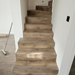 Michał Winiarski - Schody wykończone panelami drewnopodobnymi z listwami antypoślizgowymi, białe ściany, widoczny fragment podłogi i szczotka. Wykończenie wnętrza w stylu nowoczesnym.