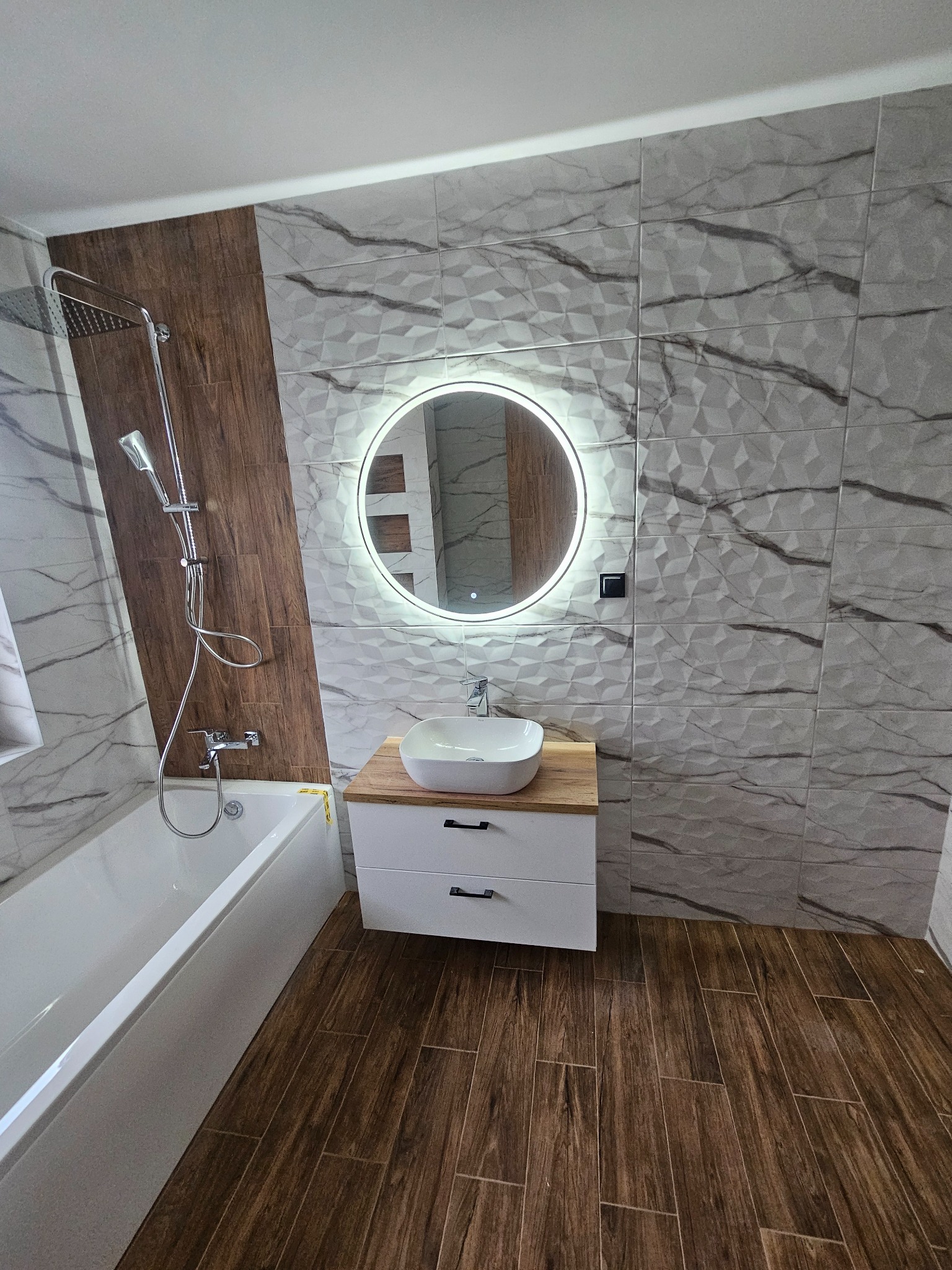 Wykończona łazienka z drewnopodobną podłogą i płytkami imitującymi marmur na ścianach, okrągłe lustro z podświetleniem LED nad umywalką nablatową.