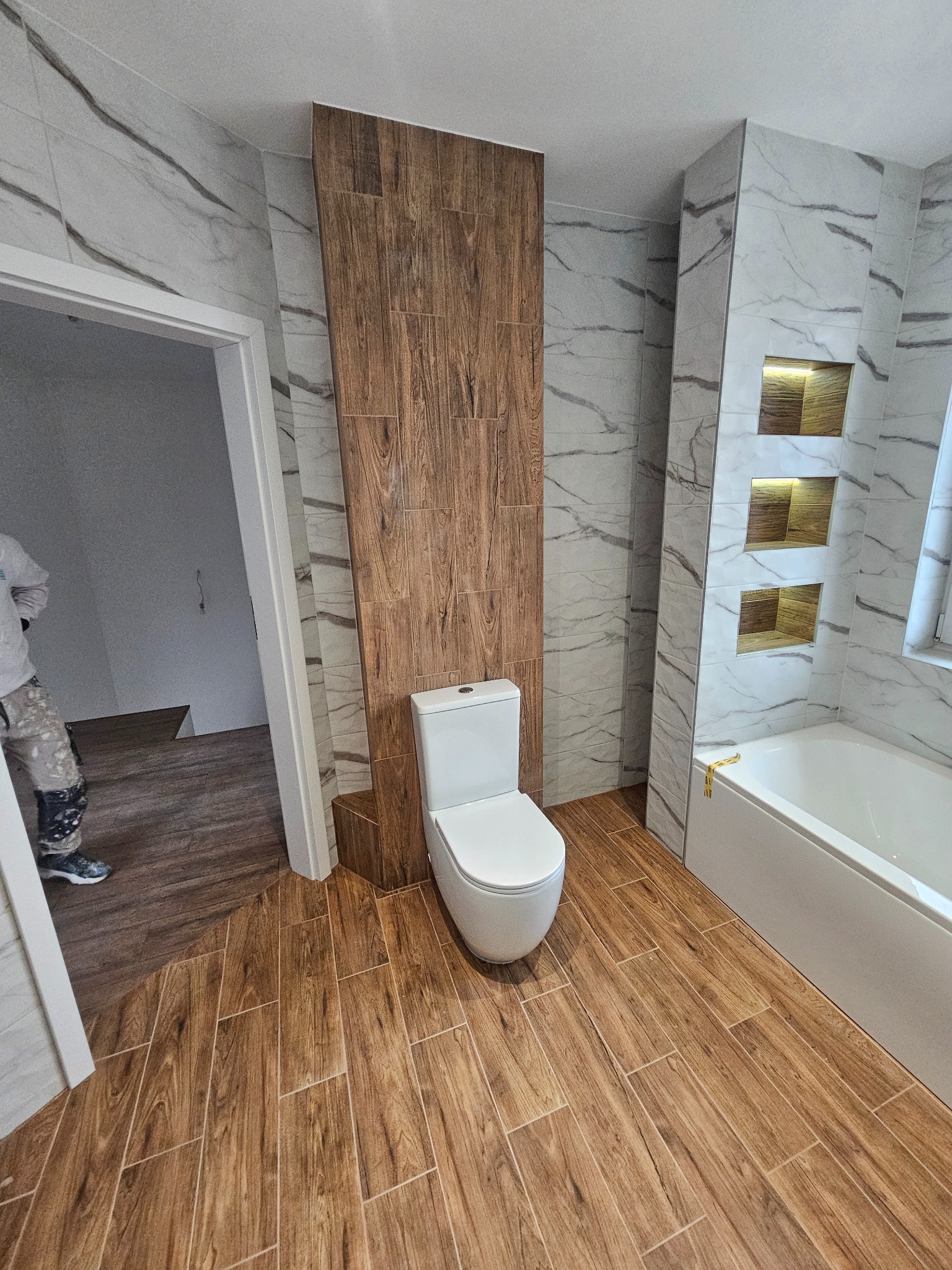 Nowoczesna łazienka z drewnopodobnymi płytkami na podłodze i ścianie, marmurowe płytki na ścianach, podświetlane wnęki obok wanny, biała toaleta i fragment osoby w roboczym ubraniu w wejściu.