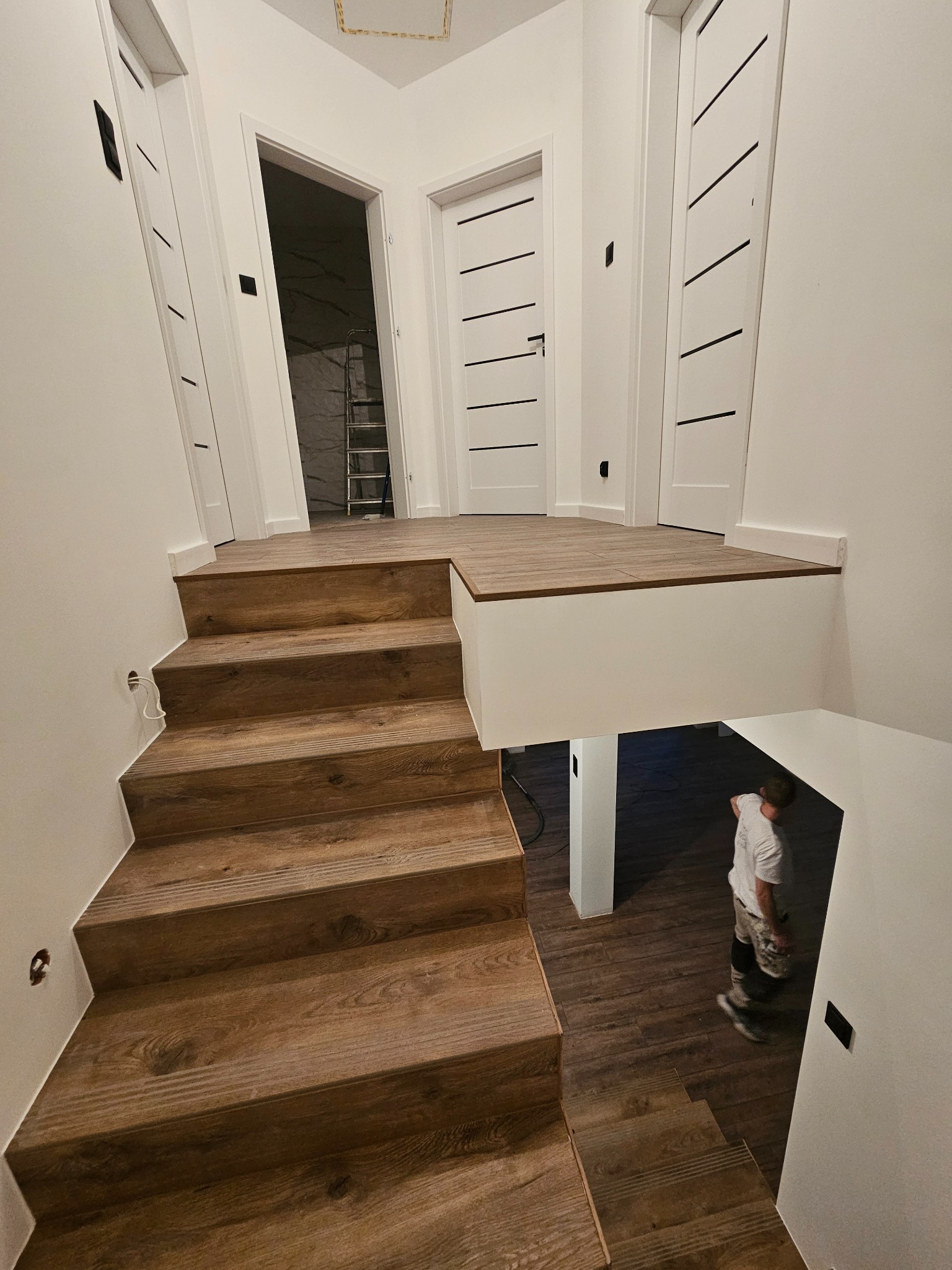 Nowoczesne schody z drewnopodobnych paneli prowadzące na antresolę, widoczny fragment osoby na dole i drabina w pomieszczeniu obok, białe ściany i drzwi z czarnymi akcentami.