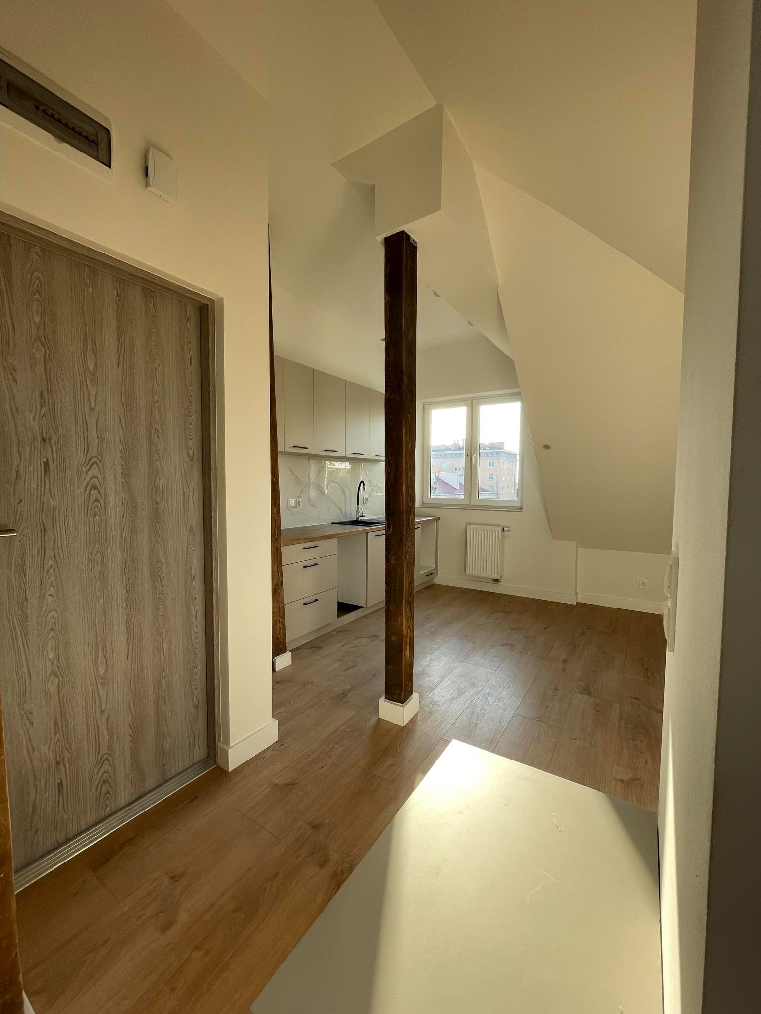 Jasne wnętrze poddasza z drewnianą podłogą i słupem konstrukcyjnym, widok na aneks kuchenny z białymi szafkami i oknem dachowym.