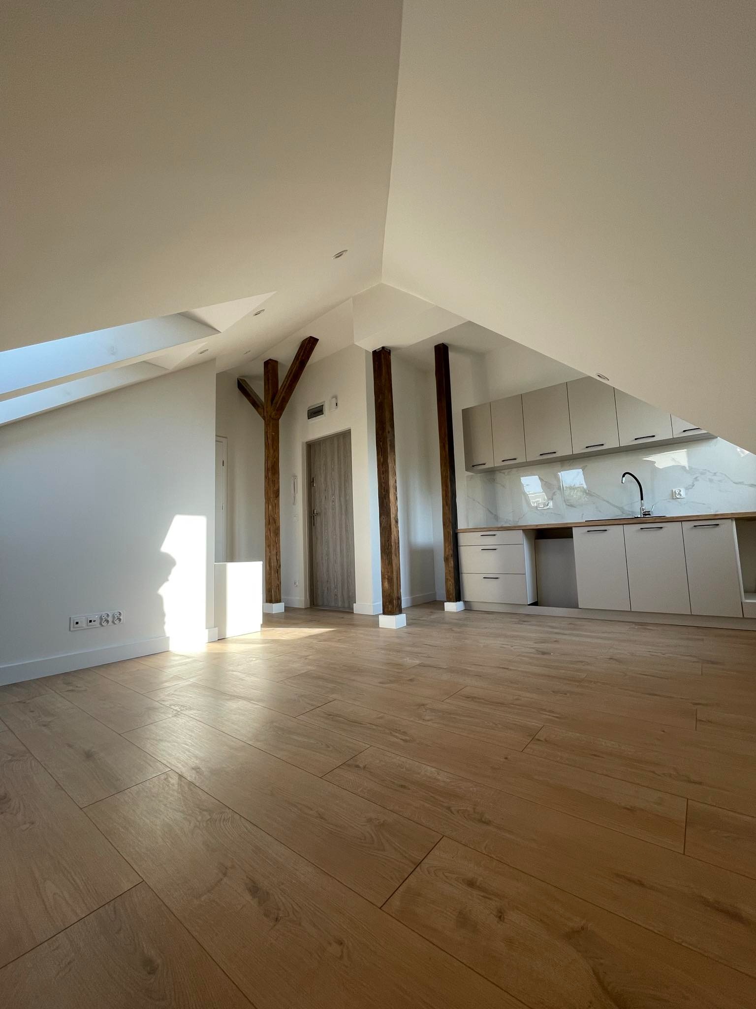 Przestronne, jasne wnętrze poddasza z drewnianymi belkami konstrukcyjnymi, nowymi panelami podłogowymi i zabudową kuchenną w odcieniach bieli, widok na okno dachowe