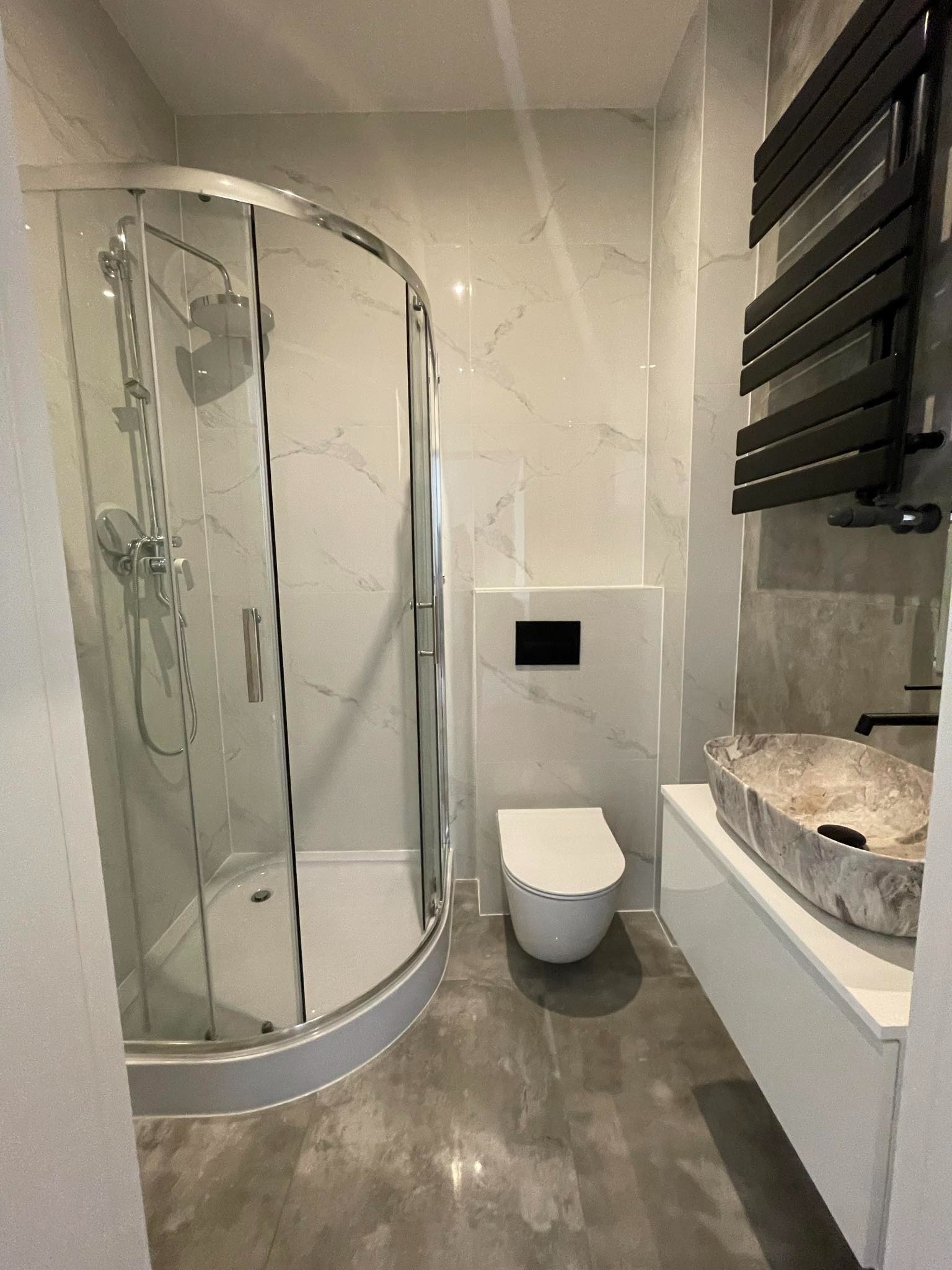 Nowoczesna łazienka z kabiną prysznicową, podwieszaną toaletą i umywalką z marmuru, wykończona płytkami imitującymi marmur, z czarnym grzejnikiem drabinkowym.
