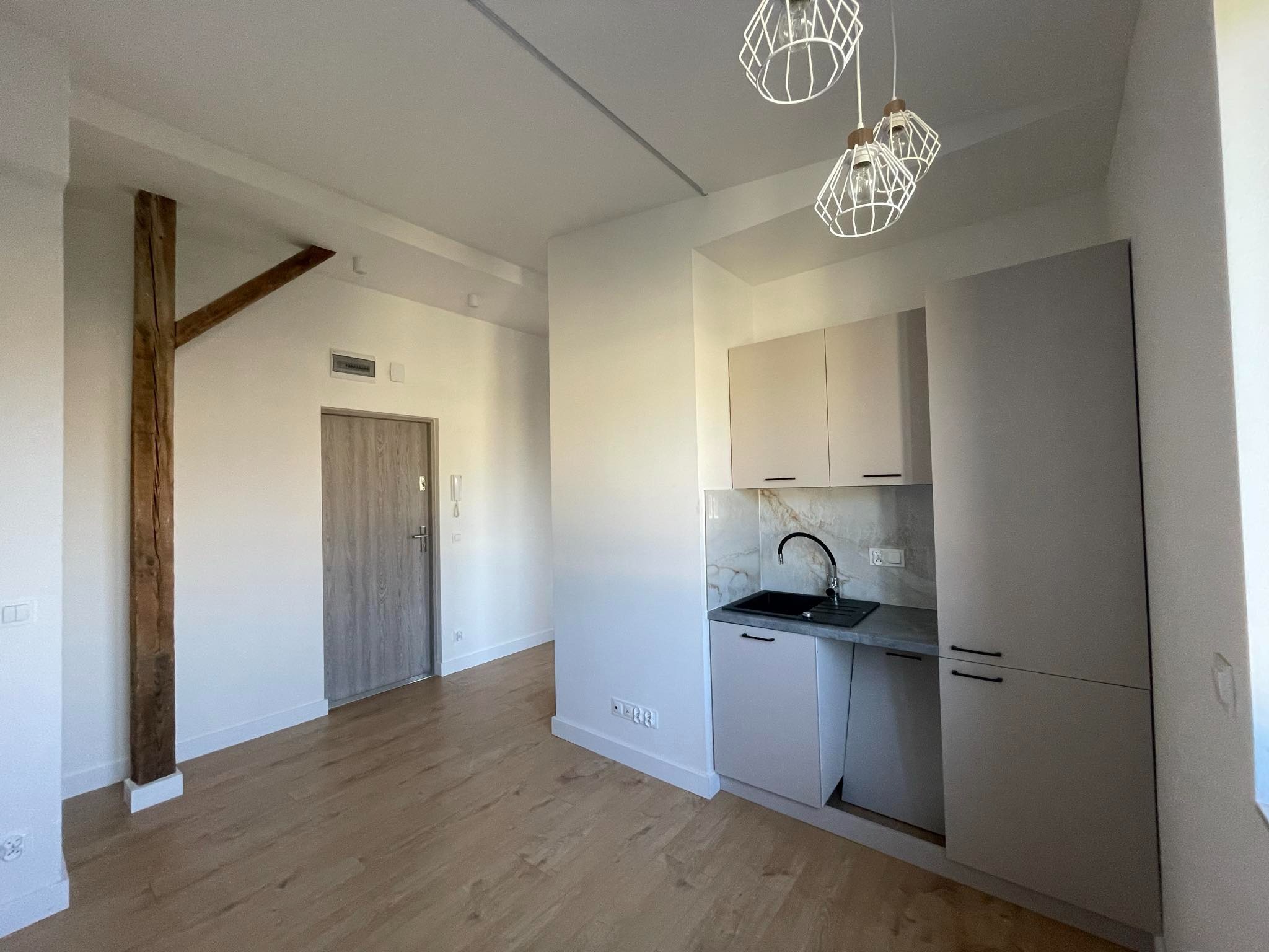 Jasne wnętrze mieszkania po remoncie, z drewnianą podłogą, minimalistyczną zabudową kuchenną w odcieniach szarości, dekoracyjnymi lampami i odsłoniętym fragmentem drewnianej konstrukcji nośnej.