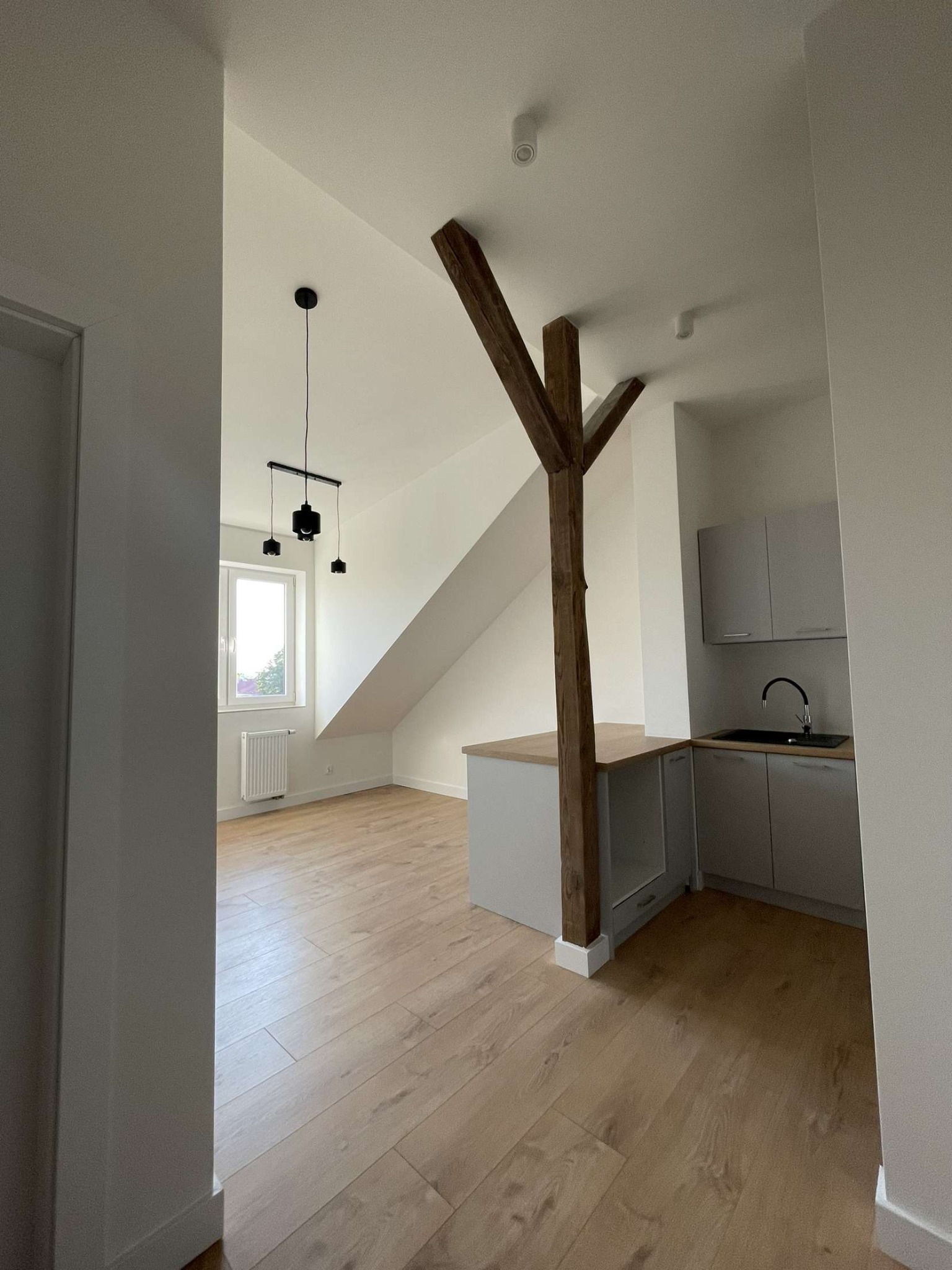 Jasne wnętrze po remoncie z drewnianą podłogą, widoczną belką konstrukcyjną, nowoczesnym oświetleniem i zabudową kuchenną w odcieniach szarości.