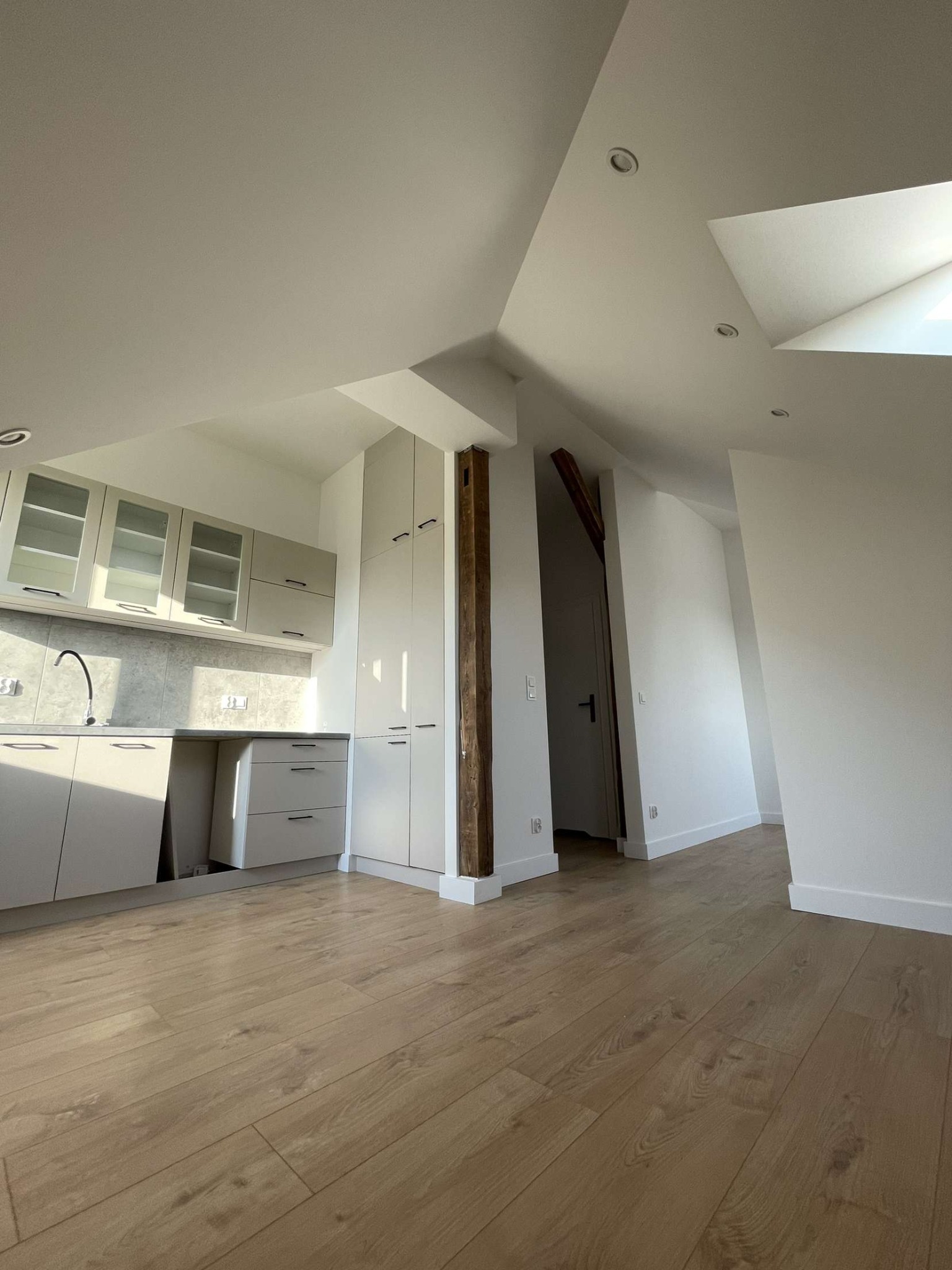 Jasna kuchnia w skosach poddasza z drewnianą podłogą, widok na fragment korytarza z drewnianą belką.