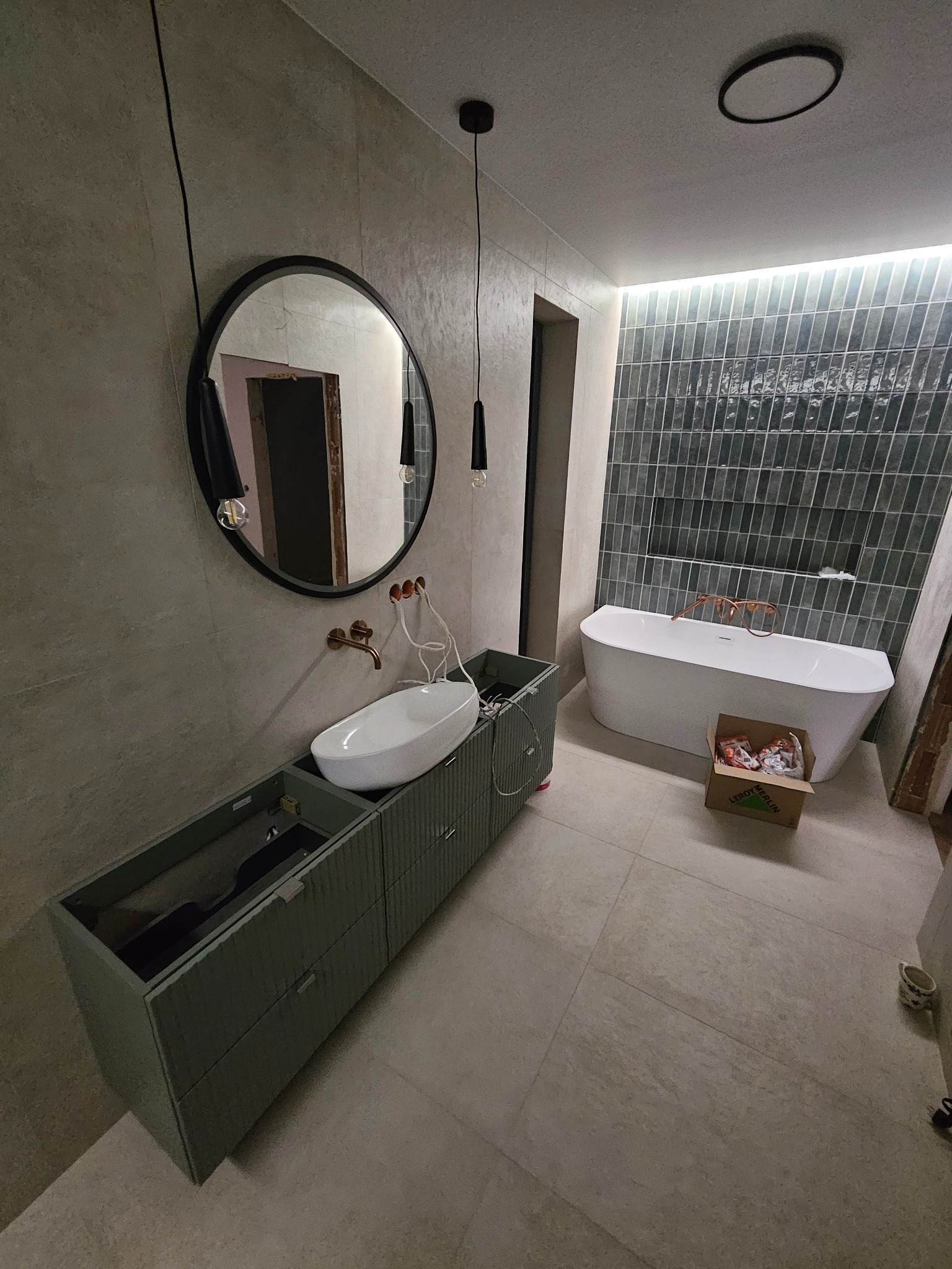 Nowoczesna łazienka w trakcie wykończenia: owalne lustro, biała umywalka nablatowa, szara szafka pod umywalkę, wanna wolnostojąca i grafitowe płytki na ścianie.