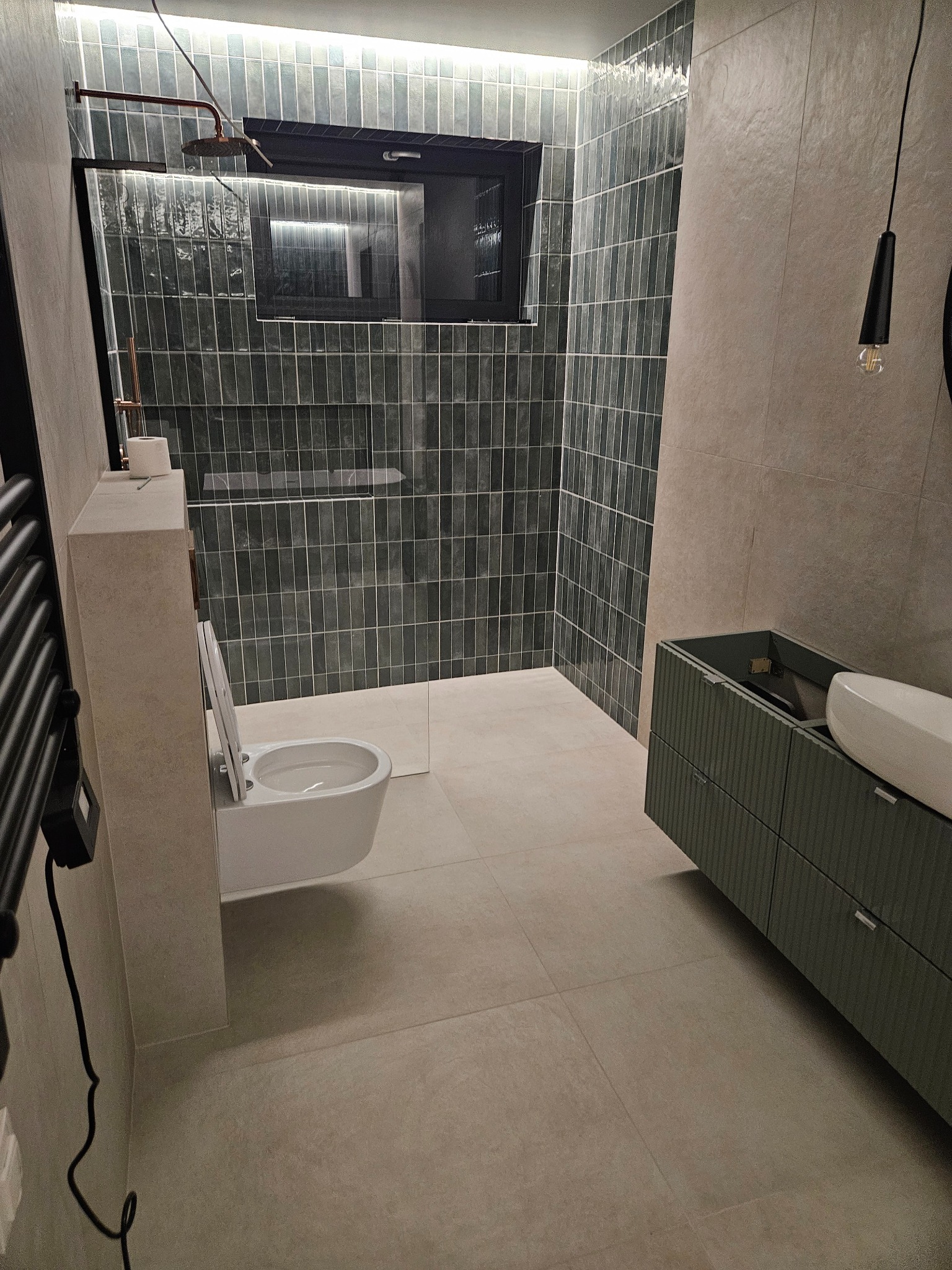 Nowoczesna łazienka z kabiną prysznicową typu walk-in, umywalką na szafce w kolorze khaki i toaletą podwieszaną. Ściany wyłożone płytkami w odcieniach szarości.