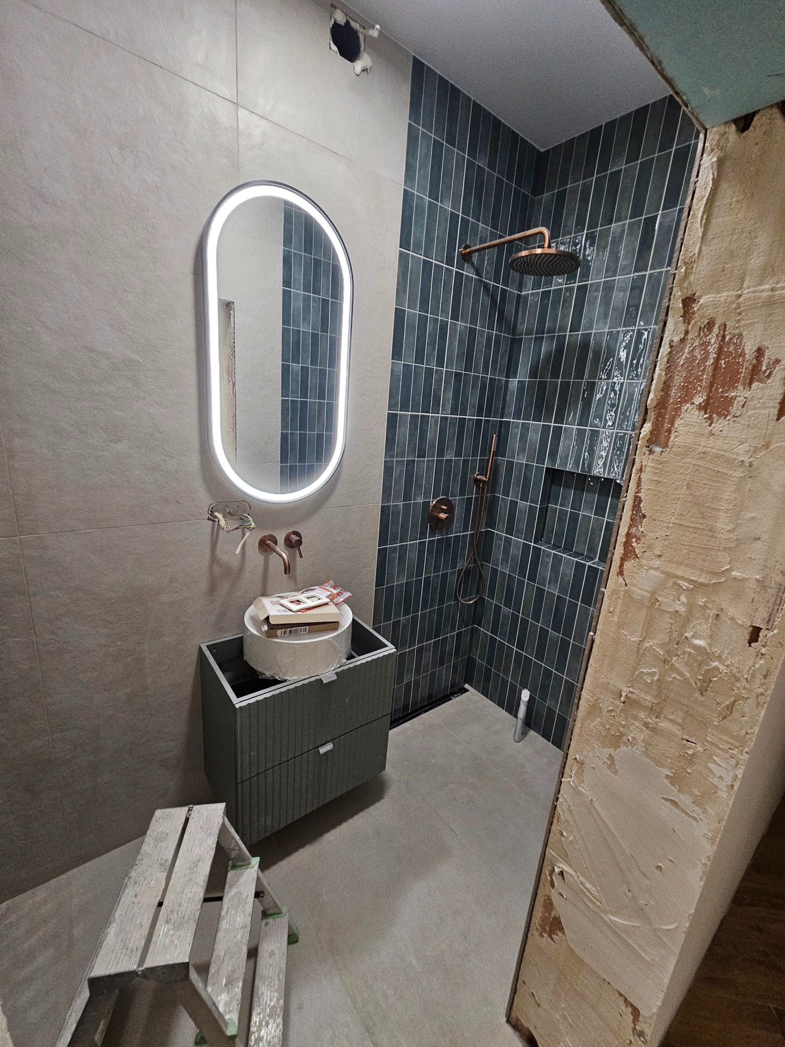 Wnętrze łazienki w trakcie wykańczania: niebieskie płytki w strefie prysznica, umywalka z szafką, lustro z podświetleniem LED i drabinka malarska.