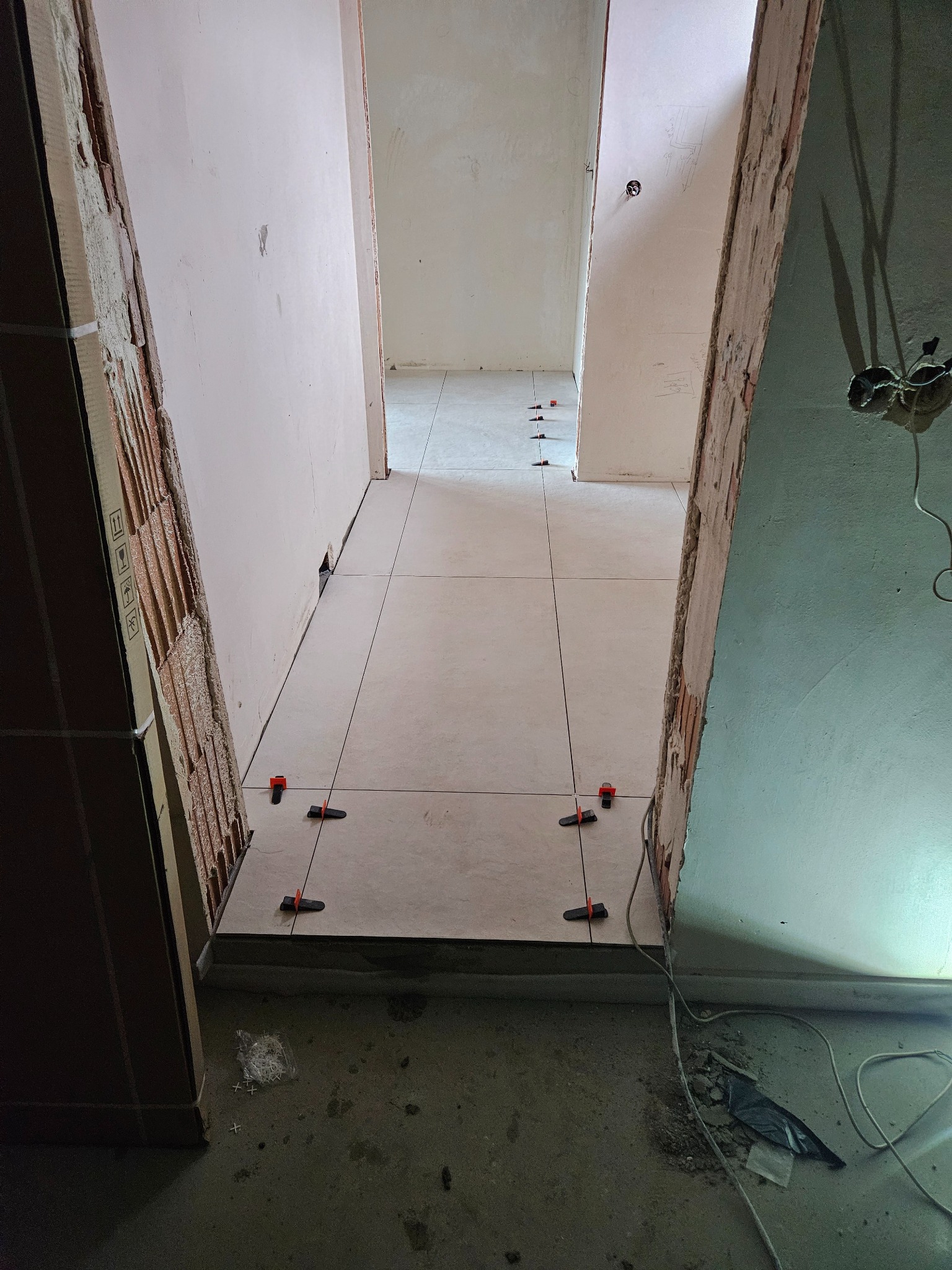 Układanie jasnych płytek podłogowych w korytarzu, z użyciem klipsów poziomujących. Widoczne surowe ściany i wystające kable elektryczne. Prace wykończeniowe w trakcie.