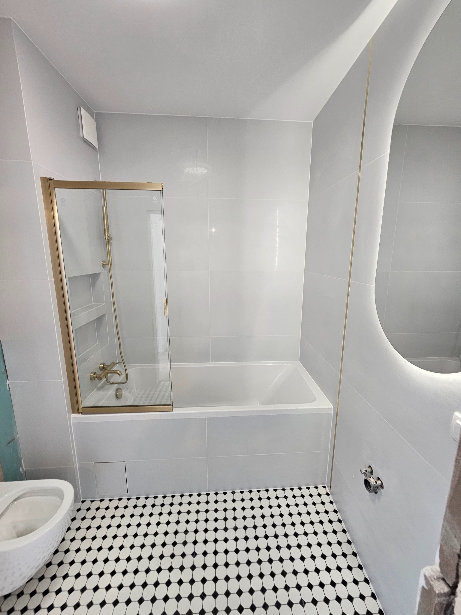 Wykończona łazienka w szarościach z wanną i kabiną prysznicową w złotej ramie. Na podłodze biało-czarna mozaika. Owalne lustro z podświetleniem LED.