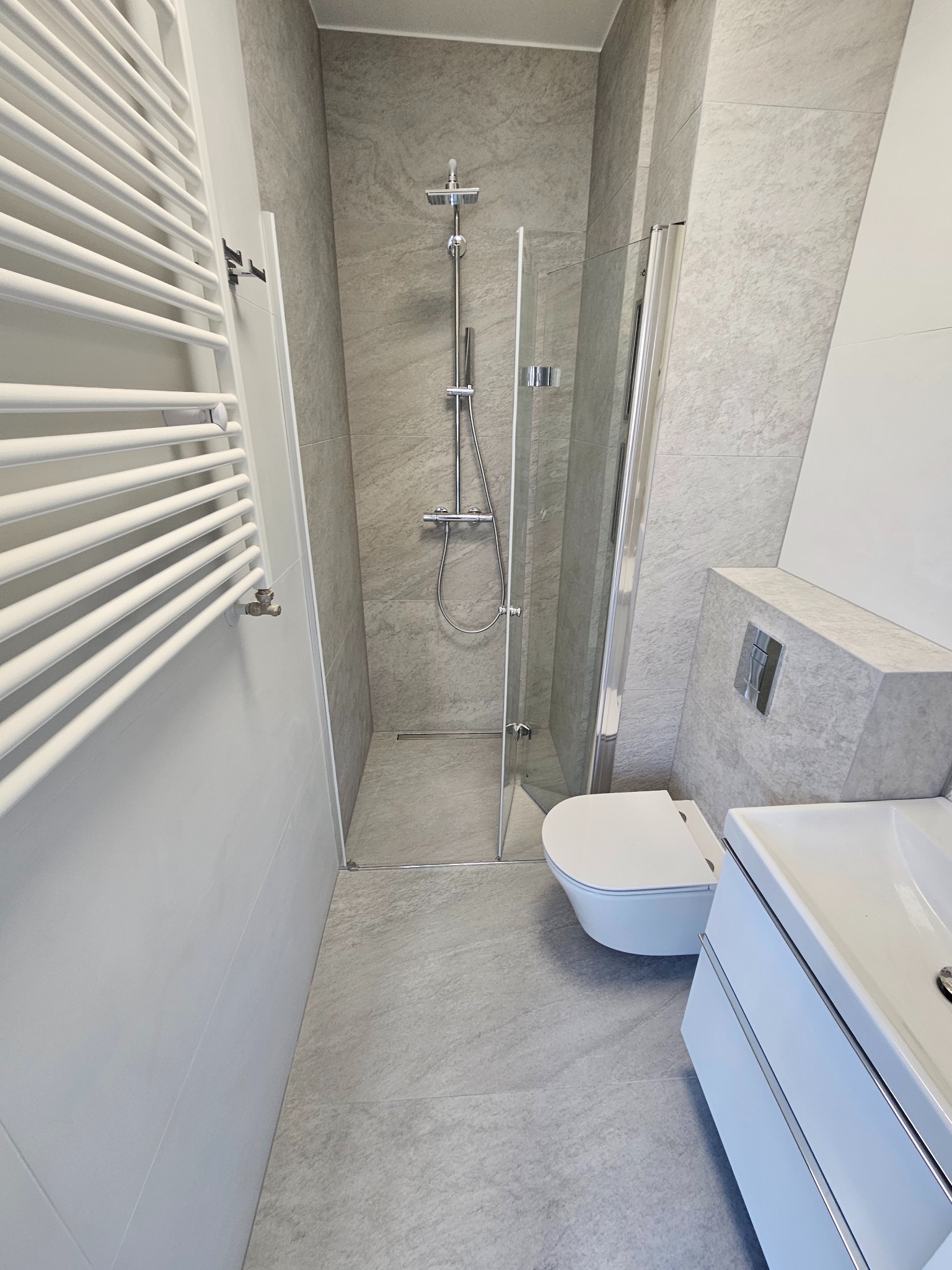 Nowoczesna łazienka w odcieniach szarości z kabiną prysznicową, wiszącą toaletą i grzejnikiem drabinkowym. Minimalistyczny design z akcentami chromu.