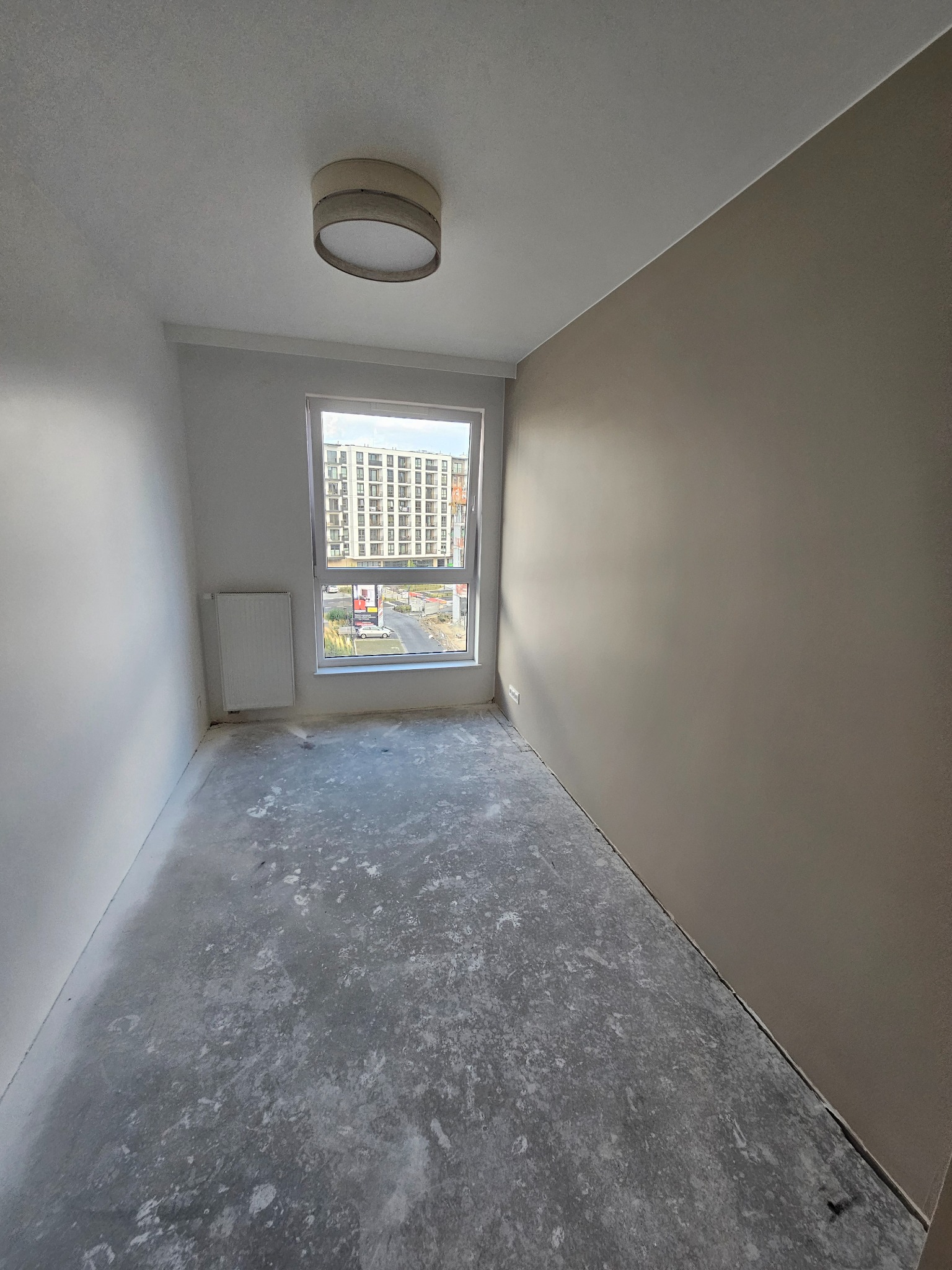 Pusty pokój po gładziowaniu ścian, z widokiem na okno i grzejnik. Podłoga surowa, przygotowana do dalszych prac wykończeniowych. Ściany w odcieniach bieli i beżu.