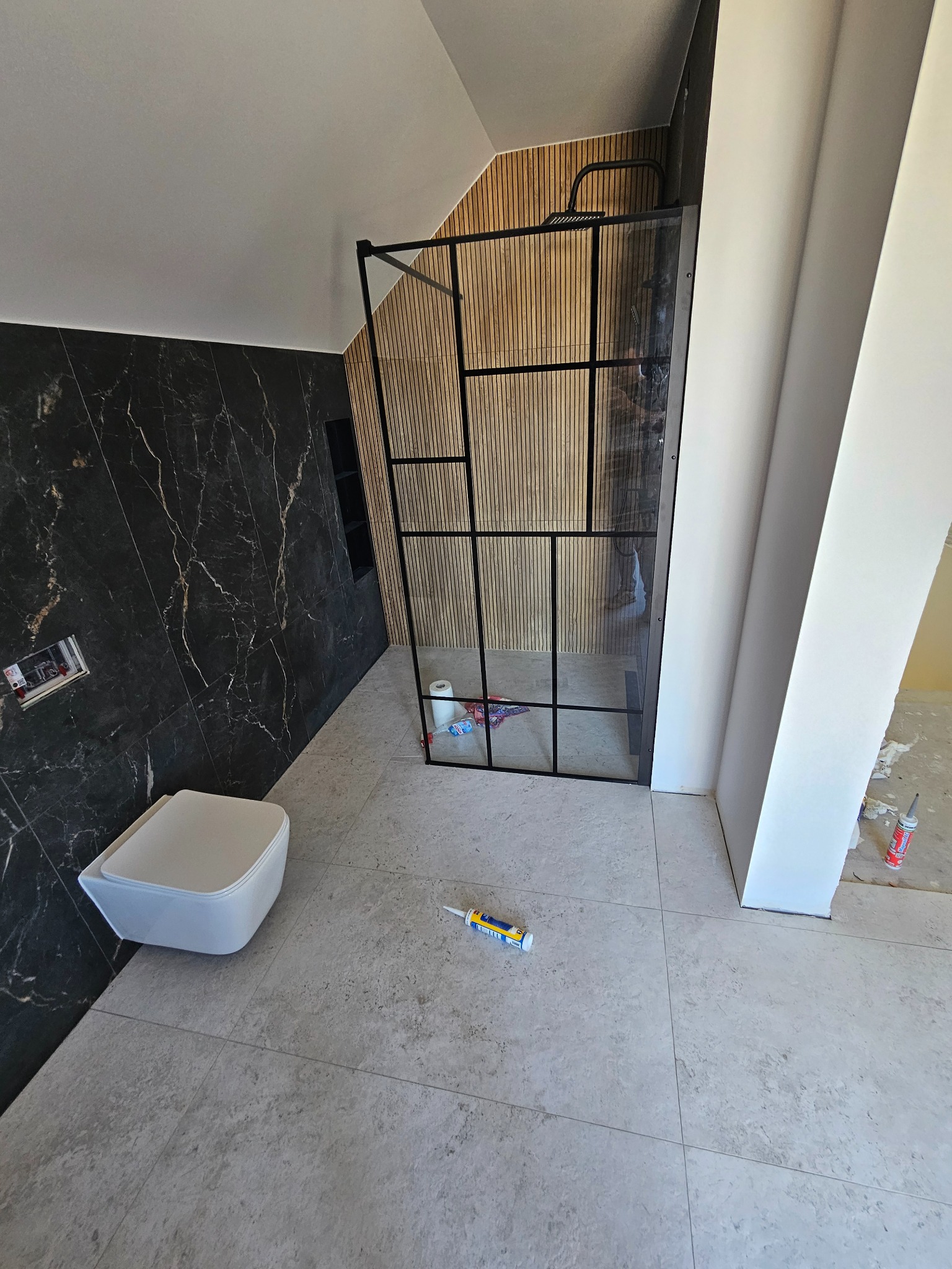 Nowoczesna łazienka w trakcie wykańczania: czarne marmurowe płytki, drewniane panele, kabina prysznicowa z czarną ramą i podwieszana toaleta. Widoczne materiały budowlane.