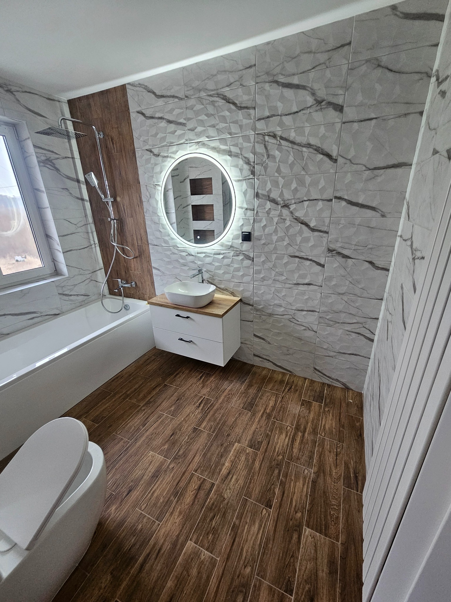 Nowoczesna łazienka po remoncie: biała wanna, umywalka z podświetlanym lustrem, płytki imitujące marmur na ścianach i drewnopodobne na podłodze.