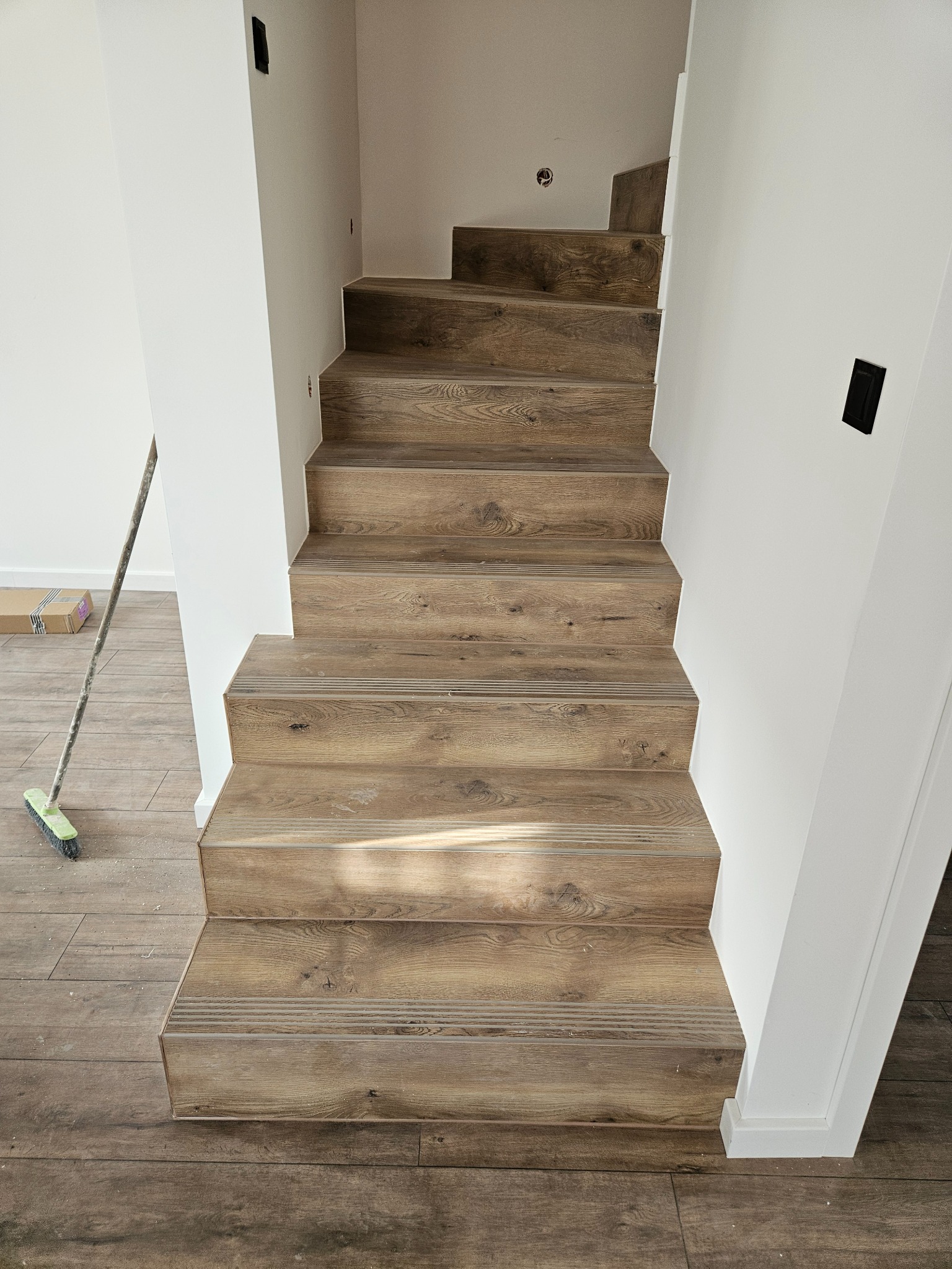 Schody wykończone panelami drewnopodobnymi z listwami antypoślizgowymi, białe ściany, widoczny fragment podłogi i szczotka. Wykończenie wnętrza w stylu nowoczesnym.