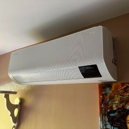 ICE TEAM Tomasz Maśluch Klimatyzacja - Biała jednostka wewnętrzna klimatyzacji Samsung WindFree zamontowana na ścianie w pomieszczeniu z beżową ścianą i obrazem olejnym w prawym dolnym rogu.