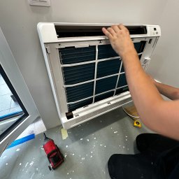 ICE TEAM Tomasz Maśluch Klimatyzacja - Instalacja białej klimatyzacji ściennej w pomieszczeniu, widoczne ręce montera, wkrętarka udarowa i miarka leżące na podłodze z plamami farby.