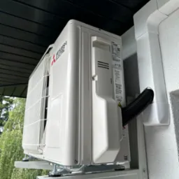 Zewnętrzna jednostka klimatyzacji Mitsubishi Electric zamontowana na wspornikach na elewacji budynku, z widocznym odprowadzeniem skroplin i fragmentem czarnego podbitki dachowej.
