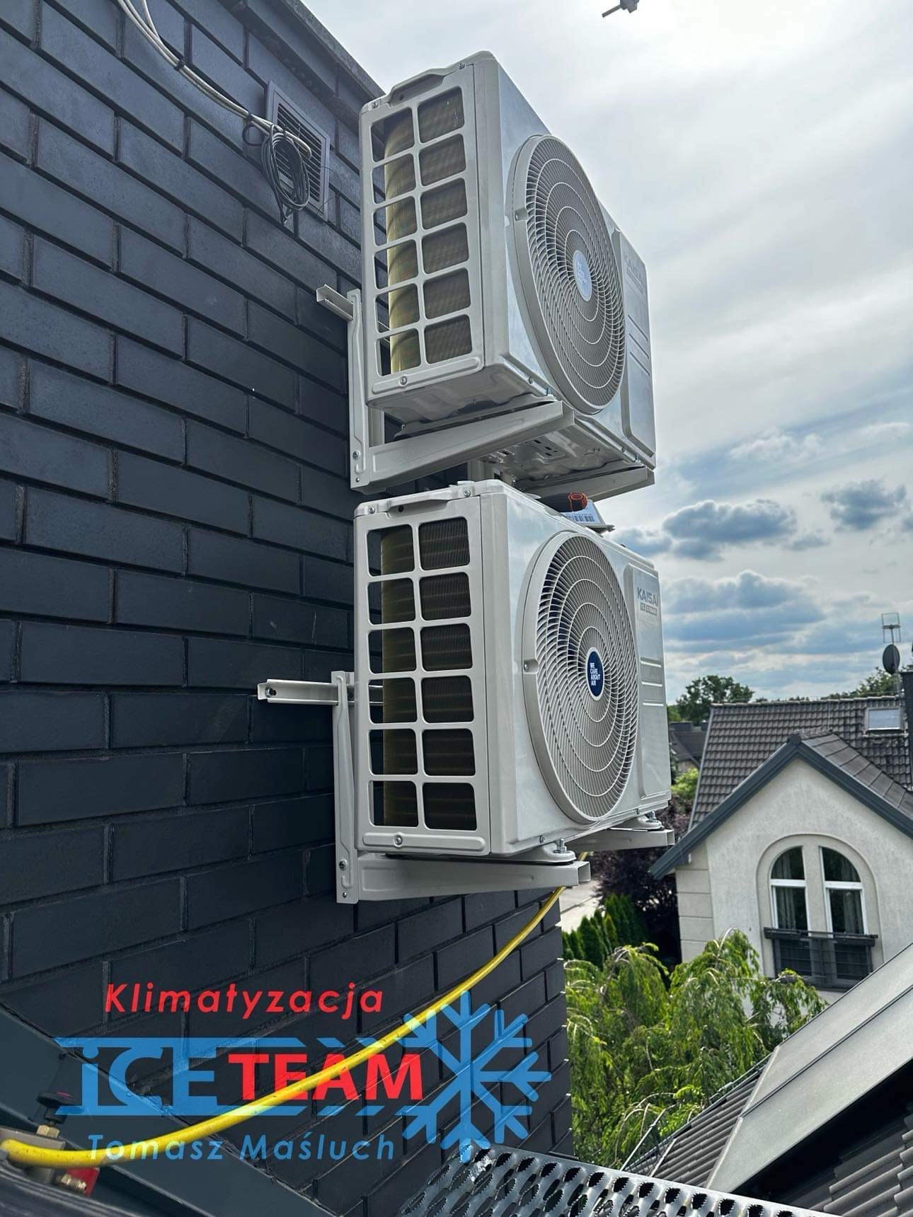 Dwa klimatyzatory typu split zamontowane jeden nad drugim na czarnej, ceglanej ścianie budynku, z widocznymi przewodami i fragmentem dachu oraz logiem firmy na dole zdjęcia.