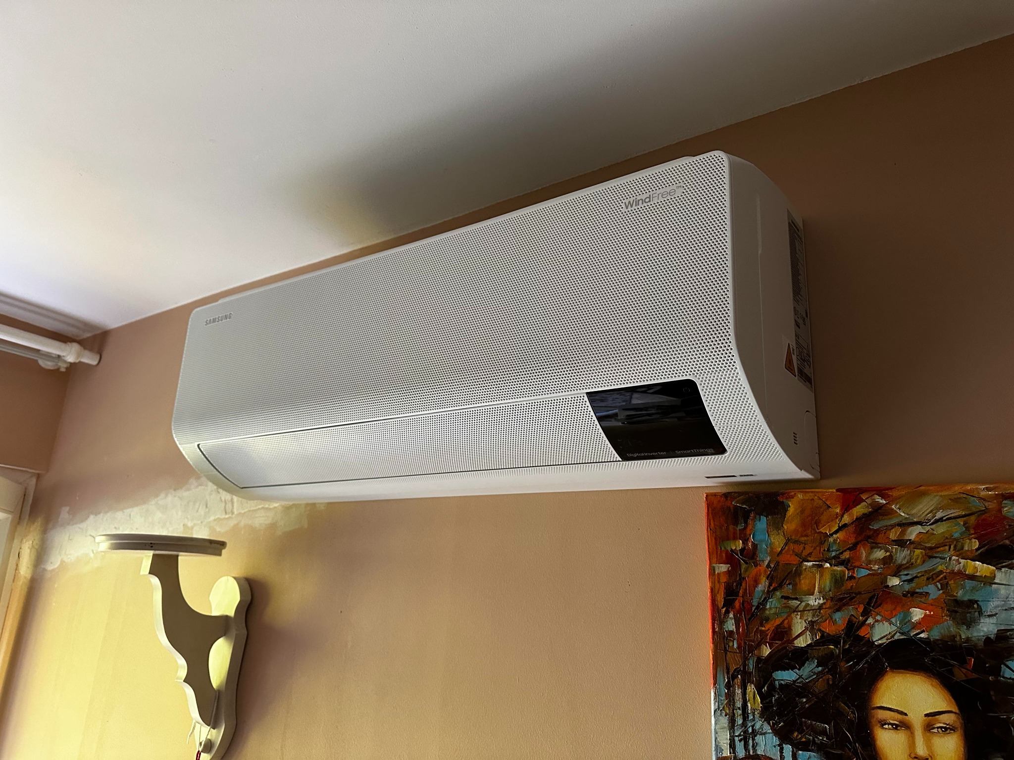 Biała jednostka wewnętrzna klimatyzacji Samsung WindFree zamontowana na ścianie w pomieszczeniu z beżową ścianą i obrazem olejnym w prawym dolnym rogu.